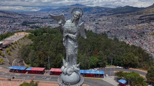 Drone begins with a close upward view of the Virgin of Quito, then pulls back to reveal the full statue, the lookout, the sprawling city below, and the surrounding mountains under cloudy sky.