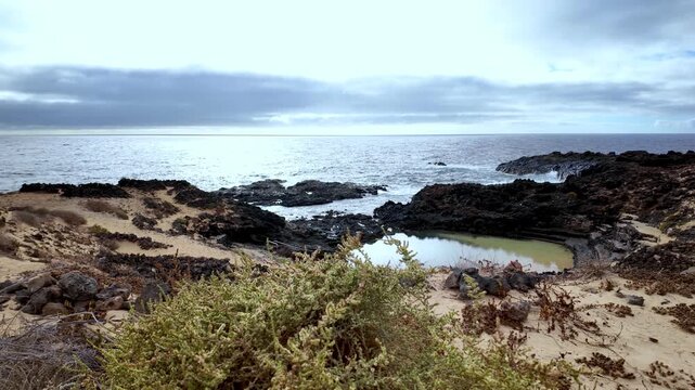 Stunning volcanic landscape featuring a natural pool at Charco del Palo, a nudist resort in Lanzarote, with the Atlantic ocean waves crashing on the rocky coast under a cloudy sky