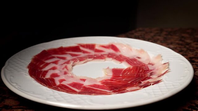 Slow motion 4K video of a professional hand arranging Iberian ham slices on a partially filled white plate. The sequence highlights the careful and artistic placement, emphasizing the marbled meat, te