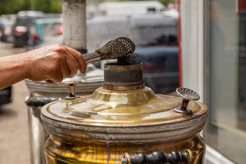 Adult hand uses tongs to place charcoal in a traditional samovar. Warm outdoor setting near building captures active, practical mood.