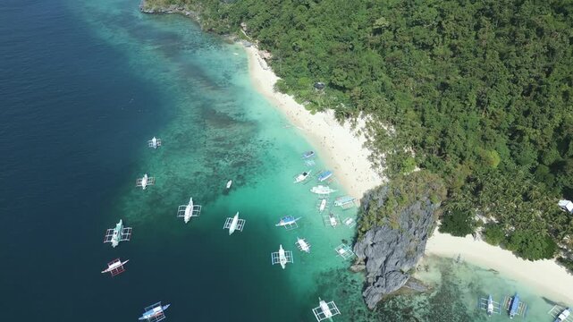 Aerial drone footage showcasing Seven Commandos Beach from a different perspective, featuring white sand, clear turquoise water, tourist boats and dramatic limestone cliffs in El Nido, Philippines.
