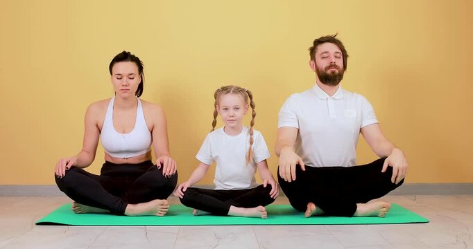 A father, mother, and daughter meditate together in the lotus position against a bright orange wall. This serene 4K footage captures mindfulness, family wellness, relaxation, and healthy lifestyle pra