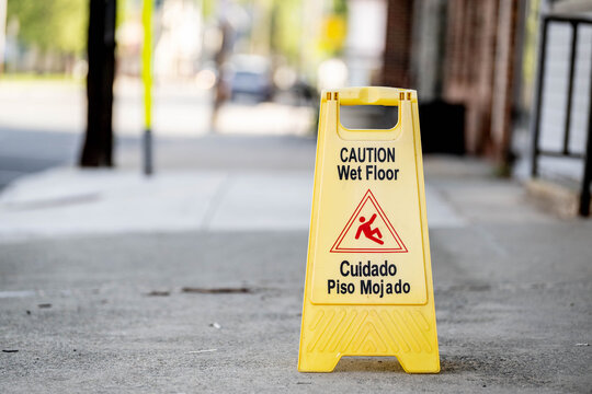 Bilingual caution wet floor sign sitting on urban sidewalk.The A-frame safety sign features English "Caution Wet Floor" and Spanish "Cuidado Piso Mojado" text, along with the universal falling person 