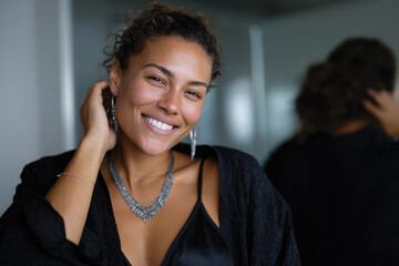 A woman smiling warmly while adjusting her hair, dressed in black with a stunning necklace. The image captures her charm and confidence in a stylish indoor setting.