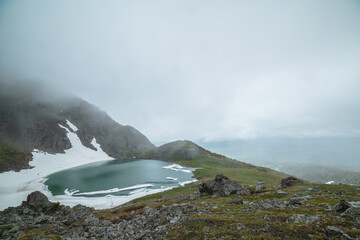 Turquoise lake and snowfield near green hill in hanging valley against misty hills in poor visibility in low clouds. Ice floes float in glacial lake in mountain cirque against foggy rainy vastness.