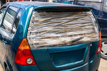 Car with damaged rear window covered with clear tape. Daytime scene reflects a temporary repair solution or poorness and inability to afford the repair.