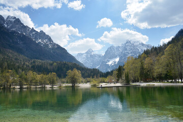 A stunning view of Jasna Lake in Slovenia with crystal clear green water reflecting the snow capped peaks of the Julian Alps under a cloudy sky
