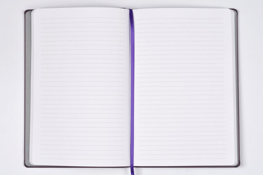 An open notebook with white lined pages and a purple silk ribbon bookmark lies flat on a neutral background featuring colored book edges