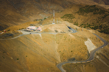 Mountain road winding through dry hills with a small roadside building and communication tower, remote highland landscape, infrastructure in rugged terrain, travel and transportation concept.