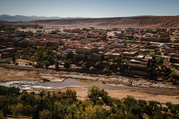 Traditional desert town with adobe houses along a dry riverbed, palm trees and oasis vegetation, surrounded by arid hills under clear sky.