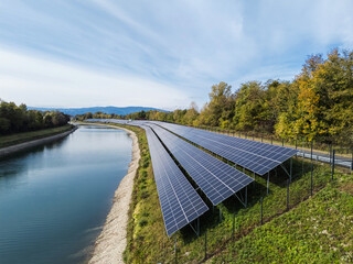 Solar panels line the bank of the river, capturing sunlight in a park setting on a clear day with trees nearby.