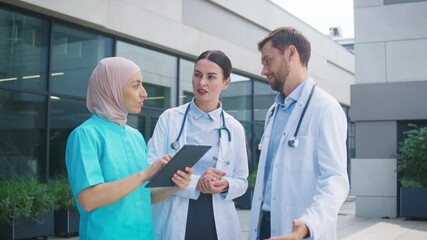 Doctors standing outside hospital. Male and female specialists listening to nurse presenting data on tablet. Team reviewing patient information calmly. Clinical setting supporting discussion. - Powered by Adobe