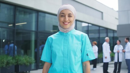 Female healthcare worker standing outside hospital courtyard wearing blue uniform. Confident professional holding steady posture and greeting viewers. Medical team exchanging updates in background.