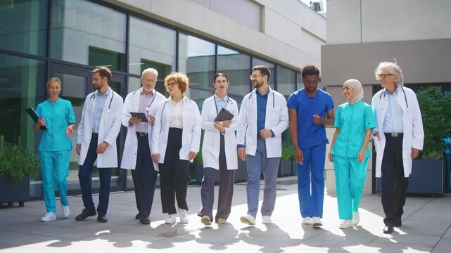 Medical team walking toward camera outside clinic showing coordinated movement. Confident doctors and nurses carrying tablets and reviewing duties while moving together. Preparing for upcoming tasks.