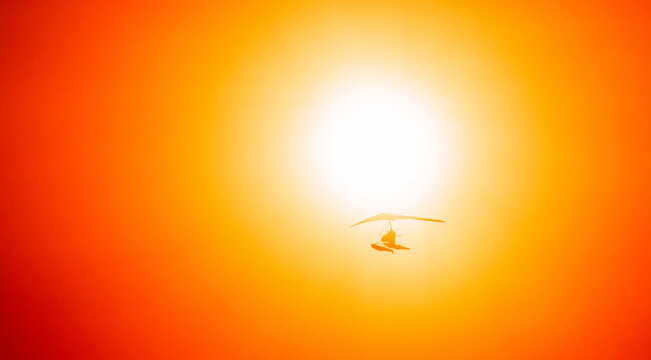 Motorized Hang Glider Flying In Clear Sunny Sunset Sky With Sun On Background