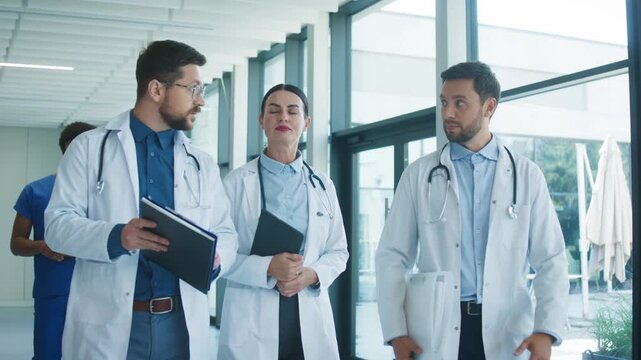 Focused medical staff walking through bright clinic hallway discussing difficult case. Serious male doctor carrying documents seeking support while colleagues listening. Team coordinating next steps.
