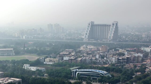 Aerial view of Greater Noida under heavy haze, showing modern high-rise offices, dense residential blocks, green patches, and a major roadway, highlighting rapid urban growth and metropolitan.