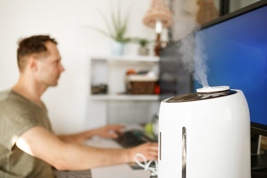 Modern air humidifier device during sitting workplace, young man using computer work on internet, enjoying aromatherapy steam scent from essential oil diffuser in room at home office.