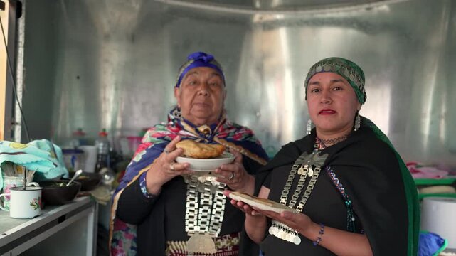 Two mapuche women wearing traditional clothes proudly show typical food in a small local kitchen