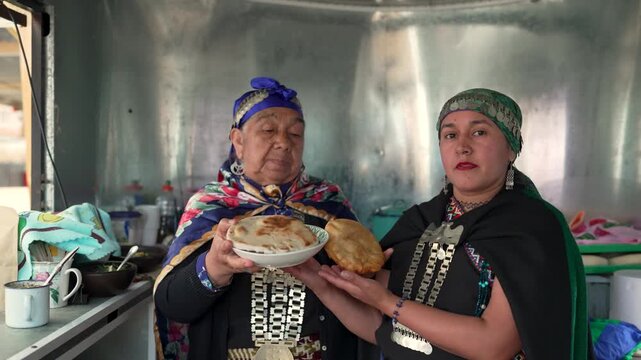 Two mapuche women in traditional clothing proudly showing and preparing typical chilean street food