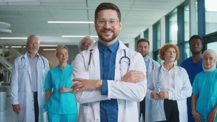 Confident Caucasian male doctor standing in bright corridor crossing arms. Smiling medical leader inspiring diverse team behind him. Showing assurance during busy hospital day. People feeling proud. - Powered by Adobe
