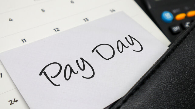 Handwritten words PAY DAY on a white note placed on a calendar with a calculator and leather folder.