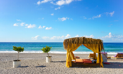 Luxury beach cabana with bright yellow curtains and thatched roof on a pebble beach. Idyllic wide view of the turquoise sea and blue sky. Exclusive summer vacation and resort concept.