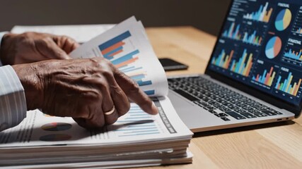Close up of an elderly african american businessman's hands flipping through financial documents with charts and graphs on a desk with a laptop showing stock market data and analytics - Powered by Adobe