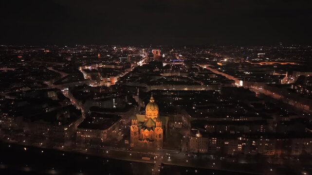 Aerial cinematography of Munich at night. The cityscape showcases the famous Frauenkirche towers and City Hall, with the riverside St. Lukas Church in the foreground under a dark evening sky