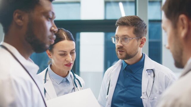 Focused doctor standing with colleagues in bright hospital room. Discussing patient update. Sharing notes and coordinating next steps during calm professional exchange. Relaxing during lunch break.