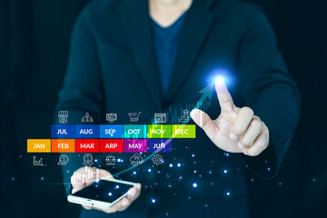 Business growth concept showing a businessman holding a smartphone and interacting with a futuristic virtual screen. The interface displays a colorful yearly calendar from January to December 