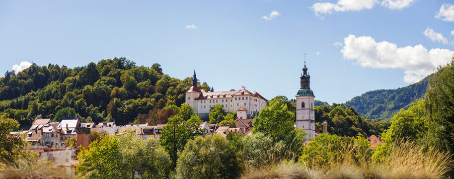 Visitors walk through the historic streets of Skofja Loka, enjoying views of medieval buildings and lush green hills on a sunny day.