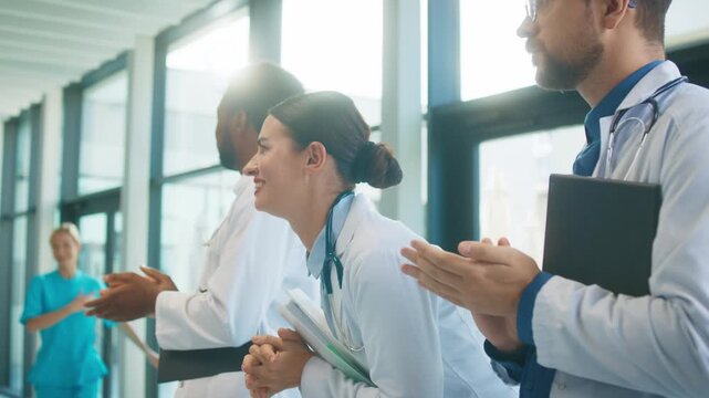Cute female doctor smiling and clapping in bright hospital corridor. Male colleagues joining applause. Team celebrating success and appreciating strong performance during busy shift.