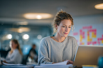 Focused young professional woman working on a laptop in a modern open-plan office, wearing a sweater with soft lighting in a collaborative workspace