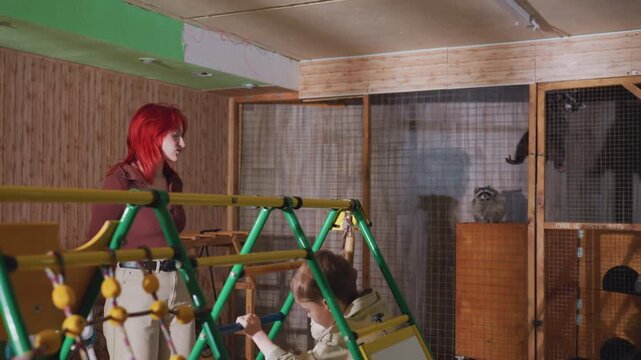 Red haired white woman inspects playset inside rustic wooden room with adjacent animal pens and cages she examines toys and structures, notes maintenance needs, and organizes cleanup as shelter
