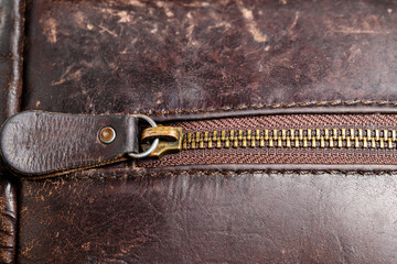 Macro shot of a brass zipper with metal teeth and leather pull tab on an aged dark brown leather bag featuring visible stitching and a textured patina surface