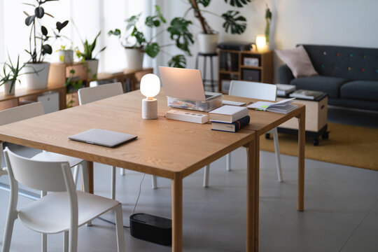 A modern home office setup with a laptop, books, and a lamp on a wooden table, surrounded by plants and minimalist furniture.