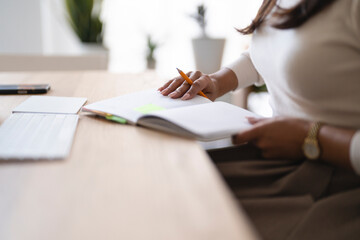 A person is writing in a notebook with a pencil, with a phone and keyboard on the desk.