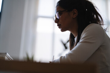 A young woman wearing glasses is focused on her work, writing with a pen at a desk.