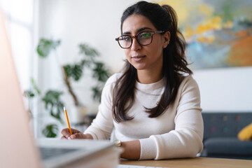A woman wearing glasses is focused on her laptop, taking notes with a pencil. She is working at a desk in a bright, modern room.