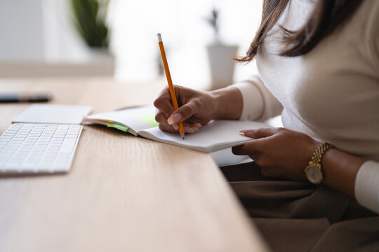 A person is writing in a notebook with a pencil at a desk with a keyboard and a plant in the background.