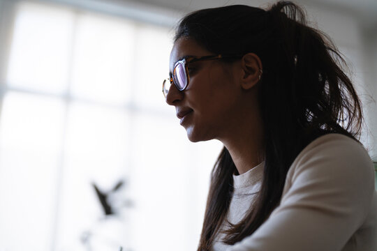 A young woman wearing glasses looks thoughtfully to the side, her hair pulled back. She is dressed in a light-colored sweater.