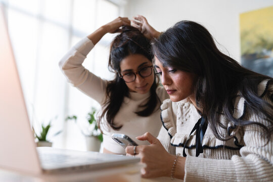 Two women are working together on a laptop and a smartphone, one is adjusting her hair.