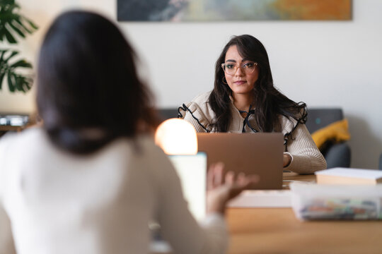 Two women are collaborating on a project at a table, one looking at a laptop while the other is engaged in conversation.
