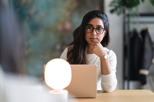 A woman wearing glasses sits at a desk with a laptop and a lamp, deep in thought as she works.
