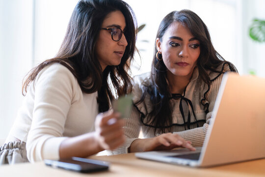 Two women collaborate on a project, one holding a card while the other types on a laptop, suggesting online shopping or data entry.