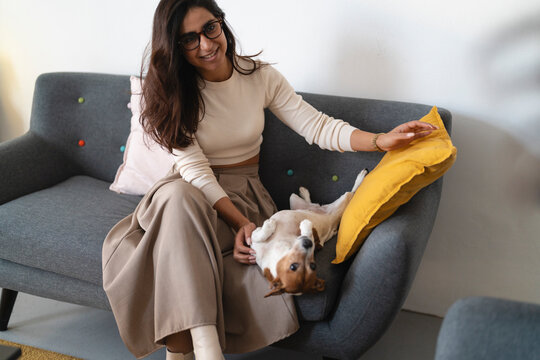 A woman with glasses smiles while petting her dog on a sofa. The dog is lying on its back, looking up at the woman.