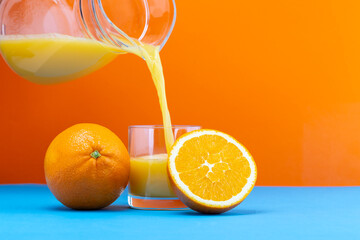 Fresh orange juice is poured into a glass with whole and sliced oranges on a blue surface against an orange background