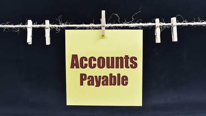 Yellow paper note with 'Accounts Payable' in dark red letters hanging on a string with wooden clothespins against a dark background