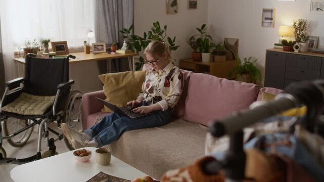 Caucasian gen Z girl with disability working on laptop while sitting on couch next to wheelchair in living room